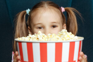 Ein Mädchen sitzt in einem Kinosessel und schaut über den Rand ihres Popcorn-Eimers.
