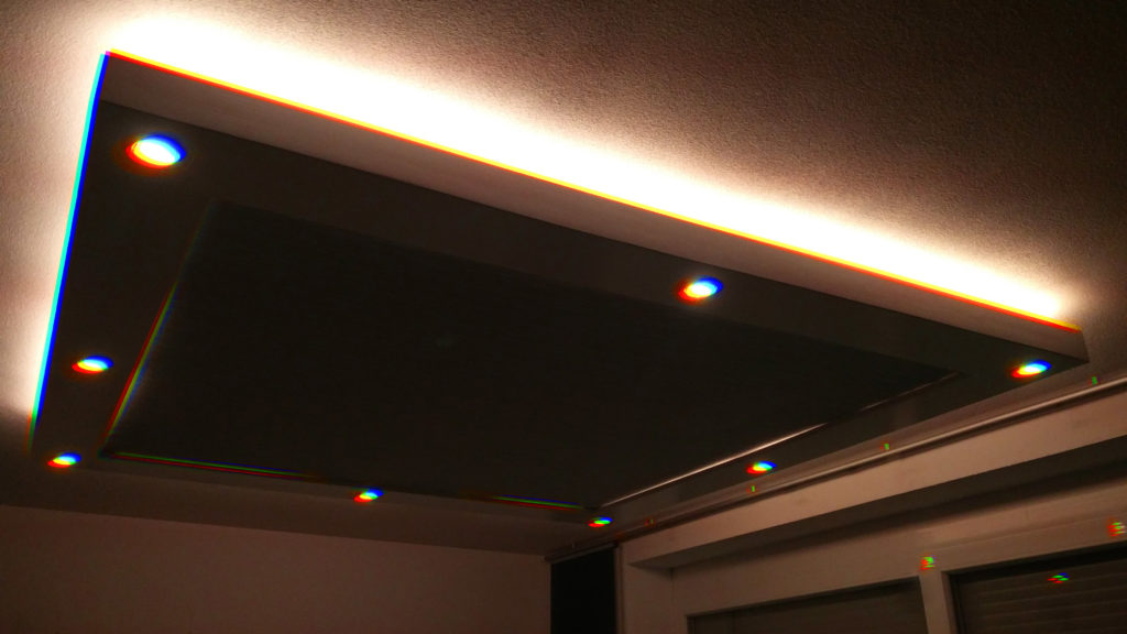 Ein Bild meines Deckensegels, überwiegend dunkler Raum mit eingeschalteten Deckenspots. Der Regenbogeneffekt wird an den hellen Spots deutlich sichtbar, die normalerweise weiß sind.